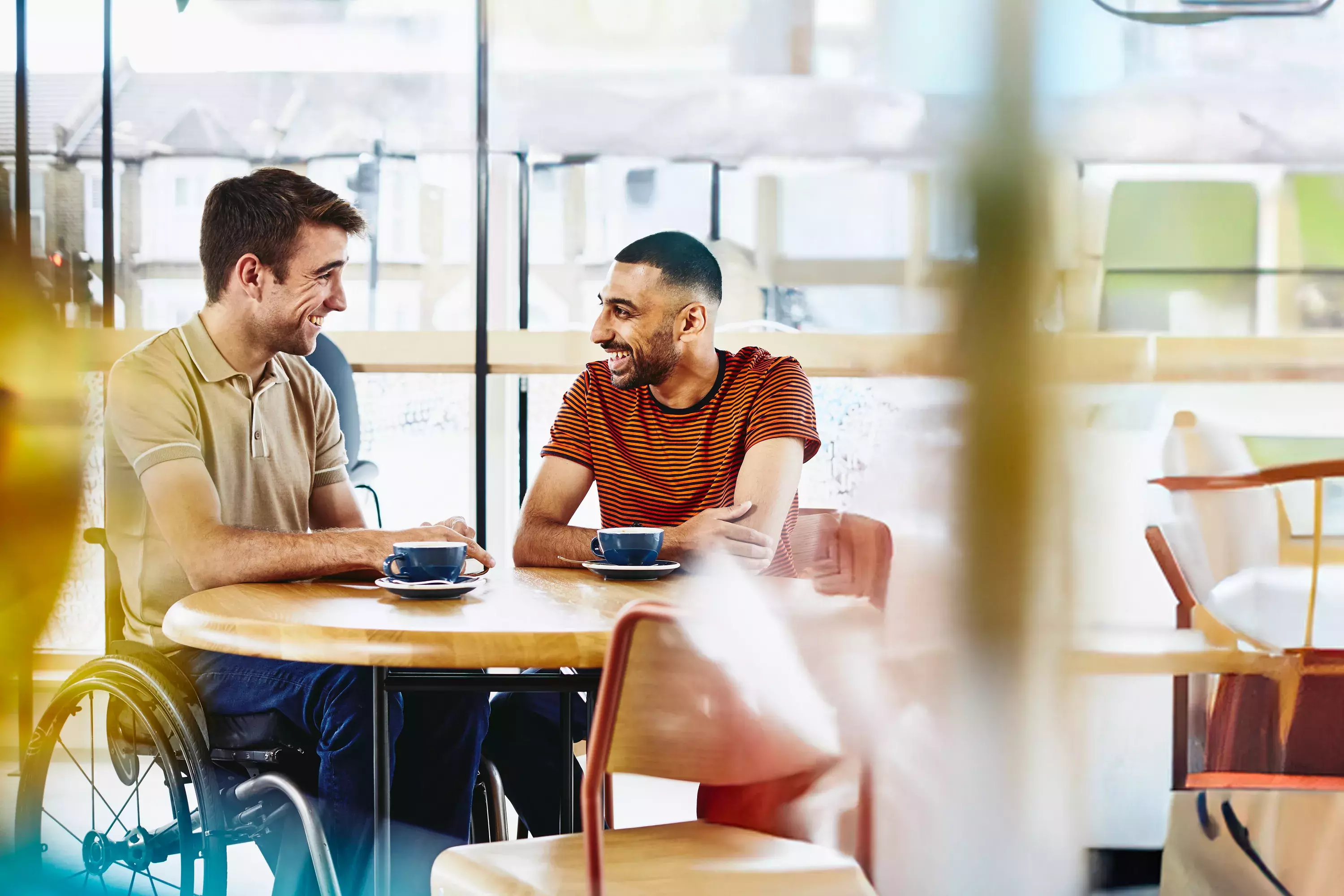 Two men sitting at a table having a conversation. Coffee cups on table.
